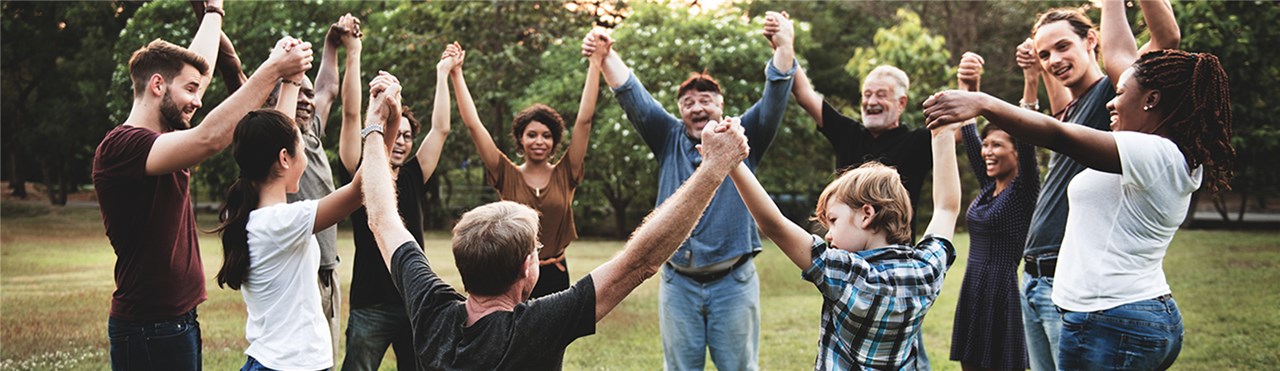 Gruppe diverser Menschen, die im Park in einem Kreis stehen und sich die Hände reichen. Es sind Menschen unterschiedlichen Alterns, Herkunft, Geschlechts sowie ein Rollstuhlfahrer dabei.