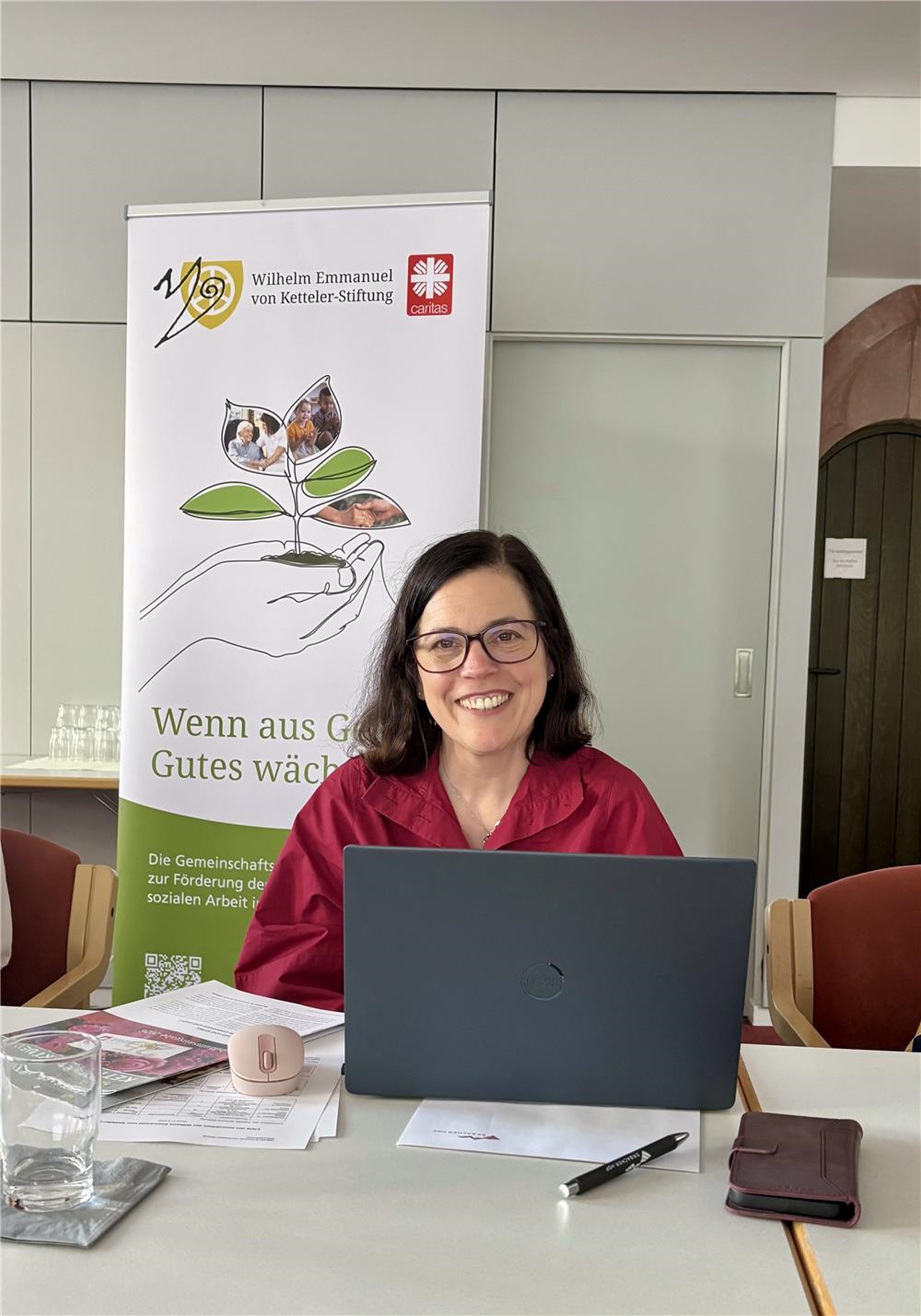 Stephanie Rieth am Laptop vor dem Roll-Up der Wilhelm Emmanuel von Ketteler-Stiftung.