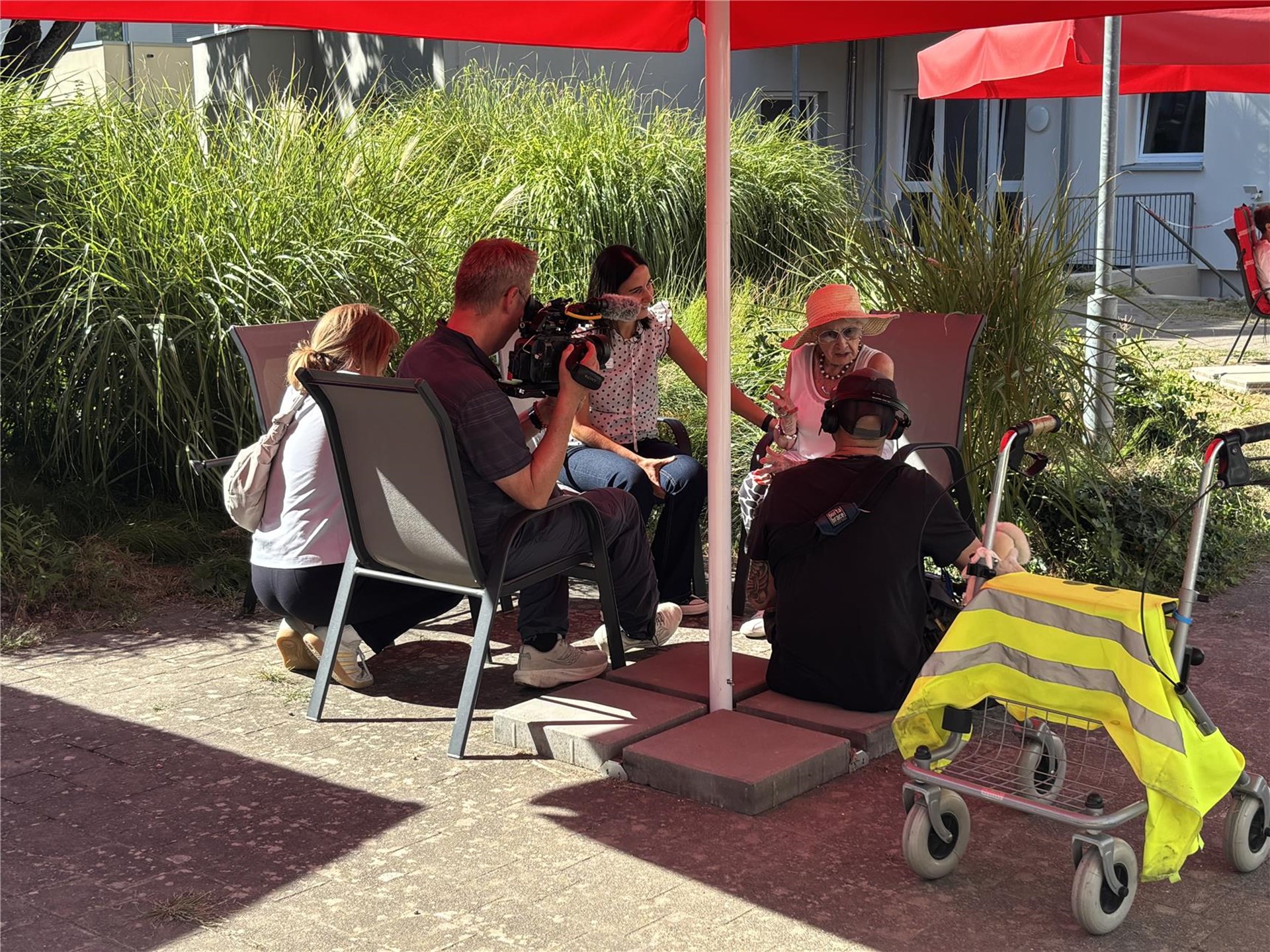 Ein Kamerateam und eine ältere Dame sitzen im Gespräch unter einem Sonnenschirm