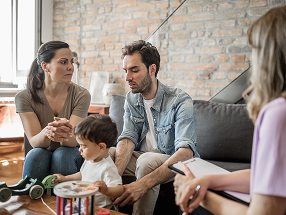 Eine Familie sitzt zusammen auf einem Sofa. Das Kind spielt mit einem Spielzeug. 