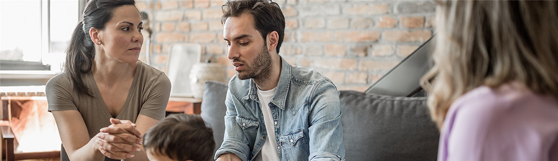 Junges Paar mit einem Kind sitzen auf einer Couch im Wohnzimmer. Mit dem Rücken zum Betrachter, aber mit Blick auf die Familie, sitzt eine Therapeutin.