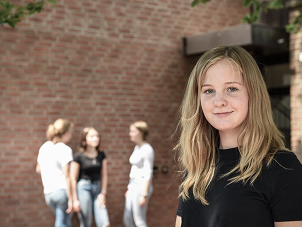 Junge Frau steht im Vordergrund. Im Hintergrund sind drei wieitere Personen unscharf zu erkennen. 