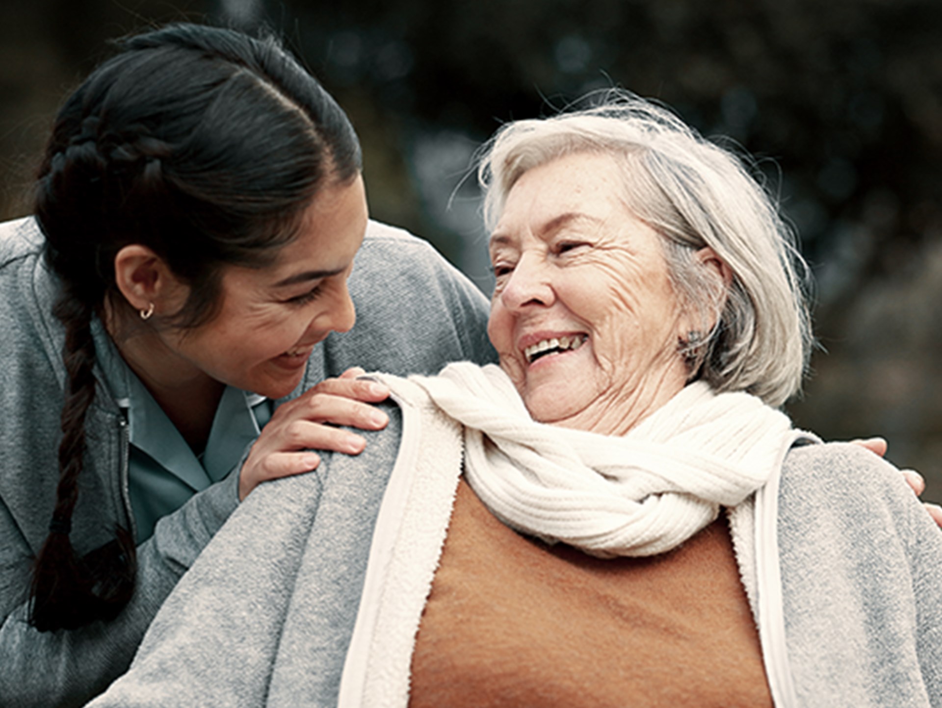 Eine Pflegekraft beugt sich zu einer einer glücklichen Seniorin im Rollstuhl und spricht mit ihr, während die beiden im Garten sind. 