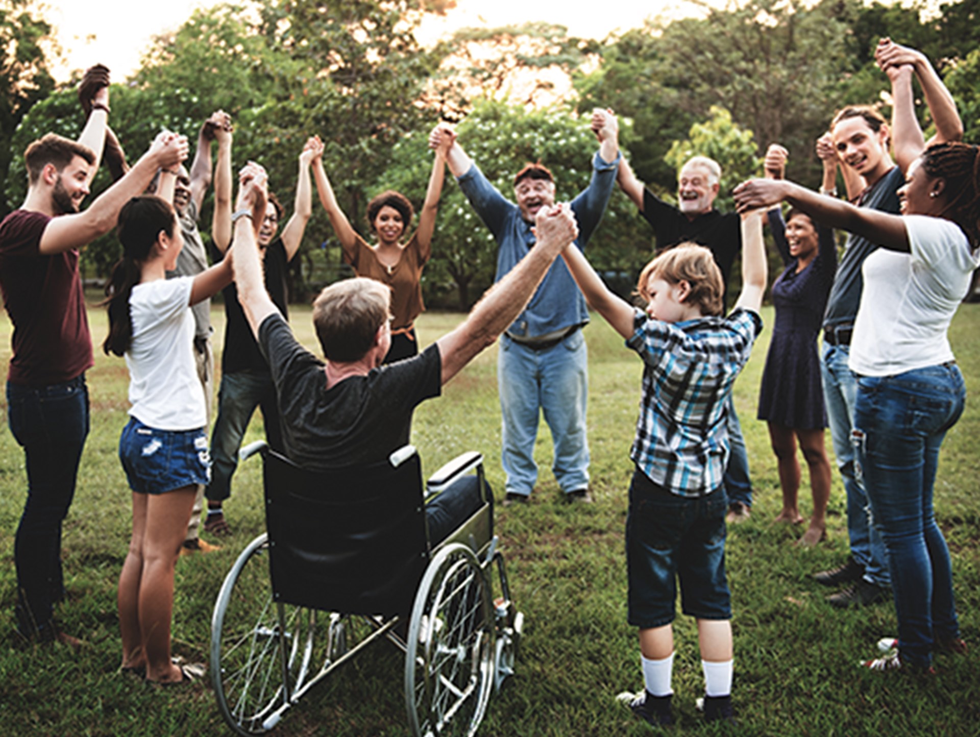 Gruppe diverser Menschen, die im Park in einem Kreis stehen und sich die Hände reichen. Es sind Menschen unterschiedlichen Alterns, Herkunft, Geschlechts sowie ein Rollstuhlfahrer dabei.